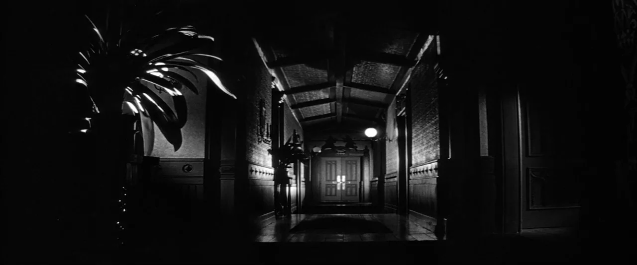 The Haunting (1963) movie still: wide — A dark, dimly lit hotel corridor features patterned walls and a central closed door. Two wall-mounted lamps…