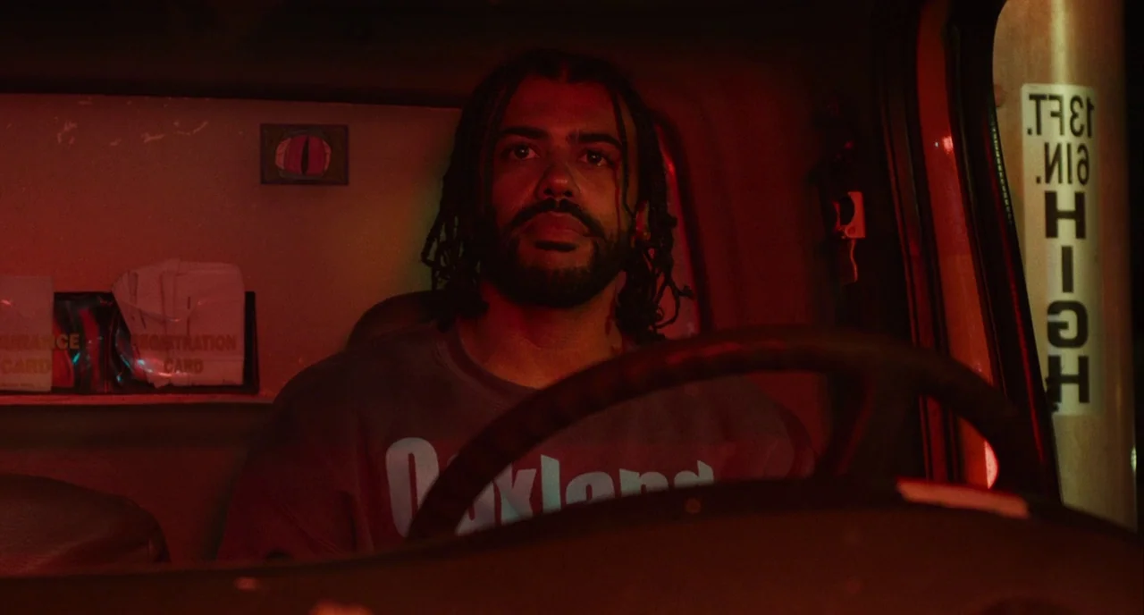 Blindspotting (2018) movie still: medium — The interior of a truck cab is illuminated by an intense red light. A man with braided hair sits at the…