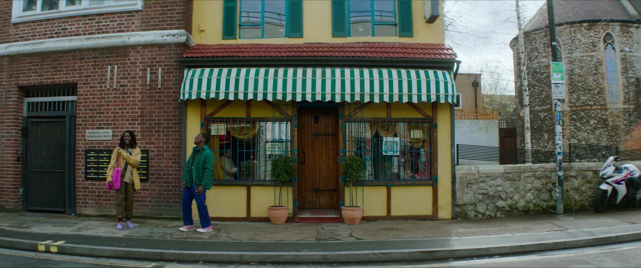 Rye Lane (2023) movie still: wide — A yellow storefront with a green striped awning sits between a brick building and stone wall. Two people stand…