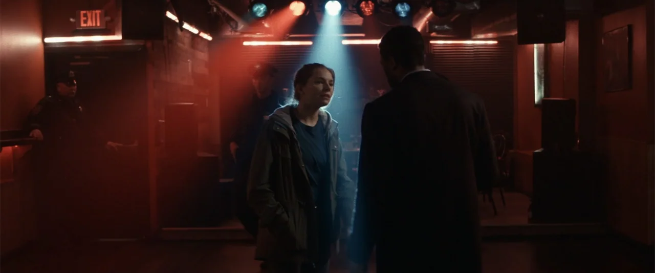 21 Bridges (2019) movie still: medium — A dimly lit nightclub interior with stage lights and shadowed figures standing on the floor. A woman speaks…