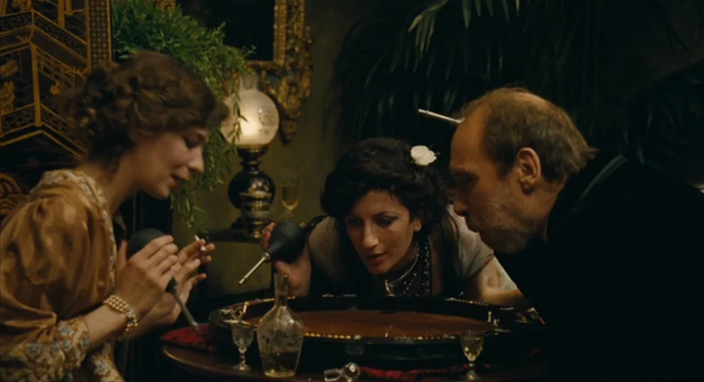 House of Tolerance (2011) movie still: medium — A dim room filled with heavy furniture and lush plants in the background. Three people sit around a…