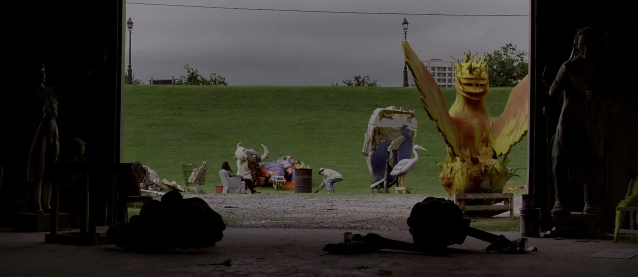 Self/less (2015) movie still: wide — A dark, cluttered industrial space opens up to a grassy field with large parade sculptures. Two figures manage…