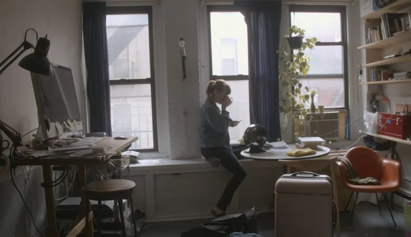 Kate Plays Christine (2016) movie still: wide — A cluttered apartment workspace features a desk, bookshelves, and a window with blue curtains. A…