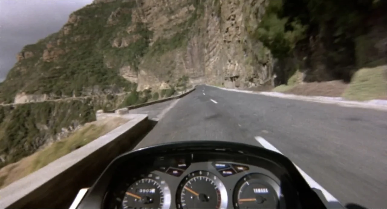 American Ninja 2: The Confrontation (1987) movie still: wide — A winding asphalt road hugs the base of a towering rocky cliffside. The viewpoint…