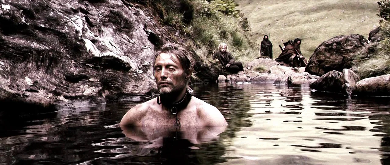 Valhalla Rising (2009) movie still: medium — A man sits neck-deep in a shallow, rocky stream surrounded by steep grassy hills. Several figures in…