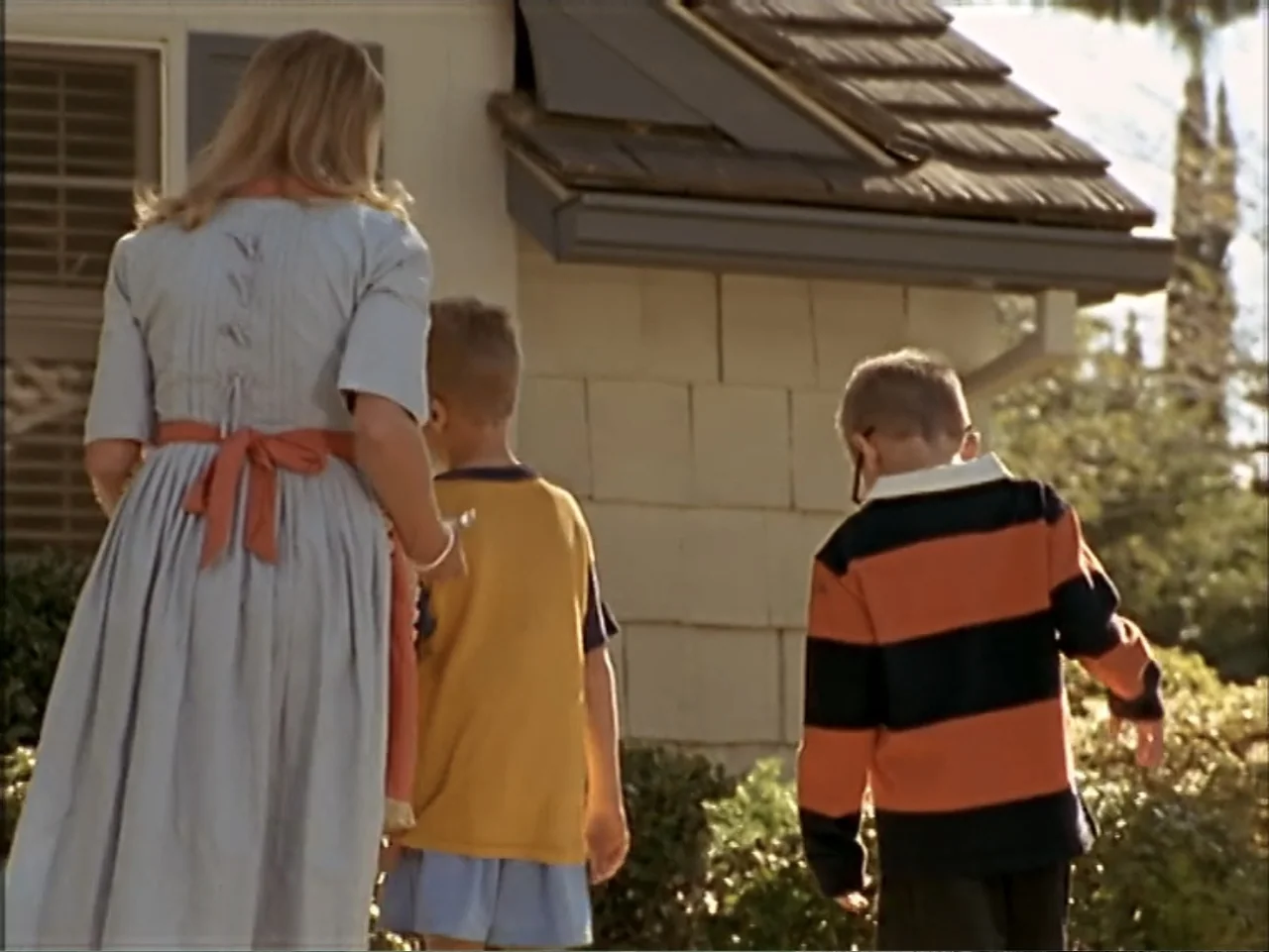 Just 4 Kicks (2003) movie still: fullbody — A tan residential home stands in the background with a sunlit front yard area. A woman and two boys walk…