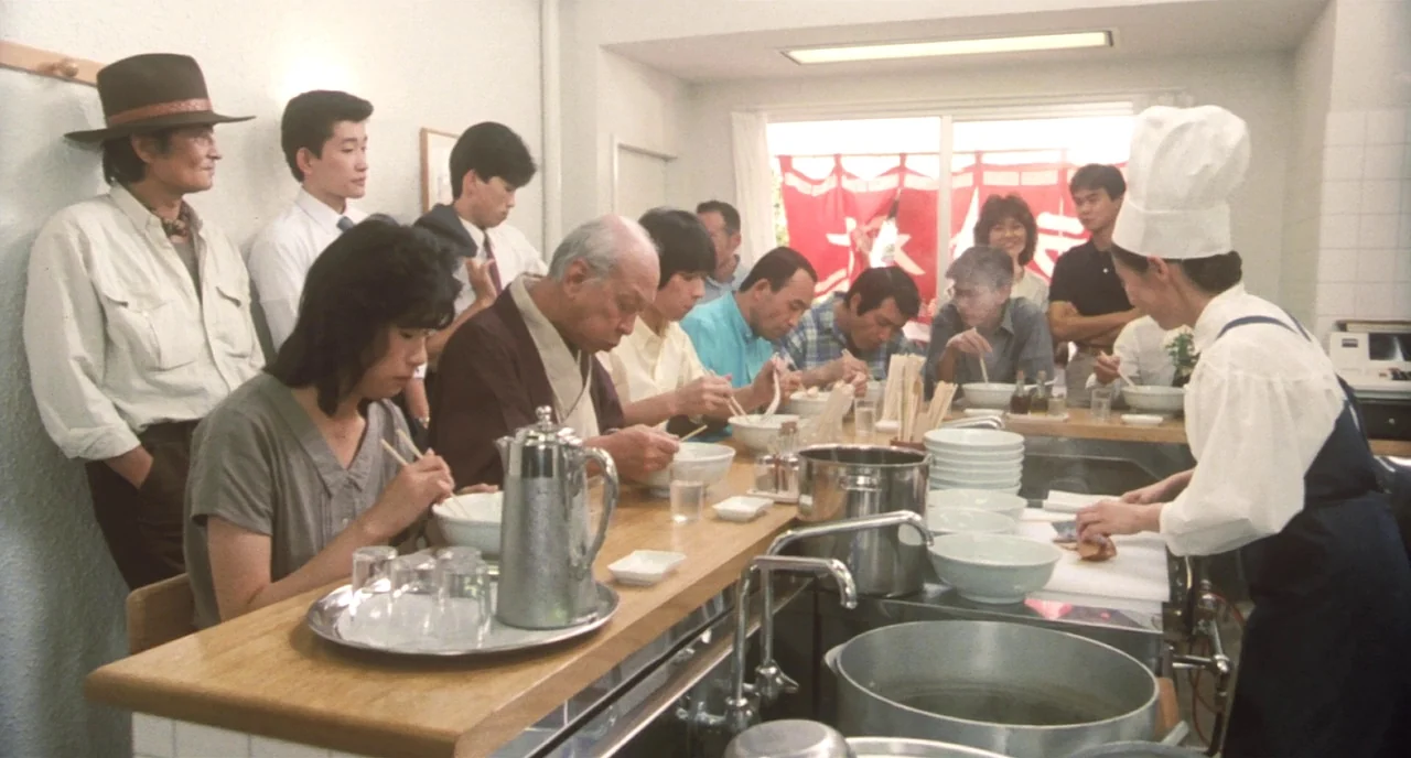 Tampopo (1985) movie still: wide — A bright, clean ramen shop features a long wooden counter and a busy preparation area. A chef in white serves…