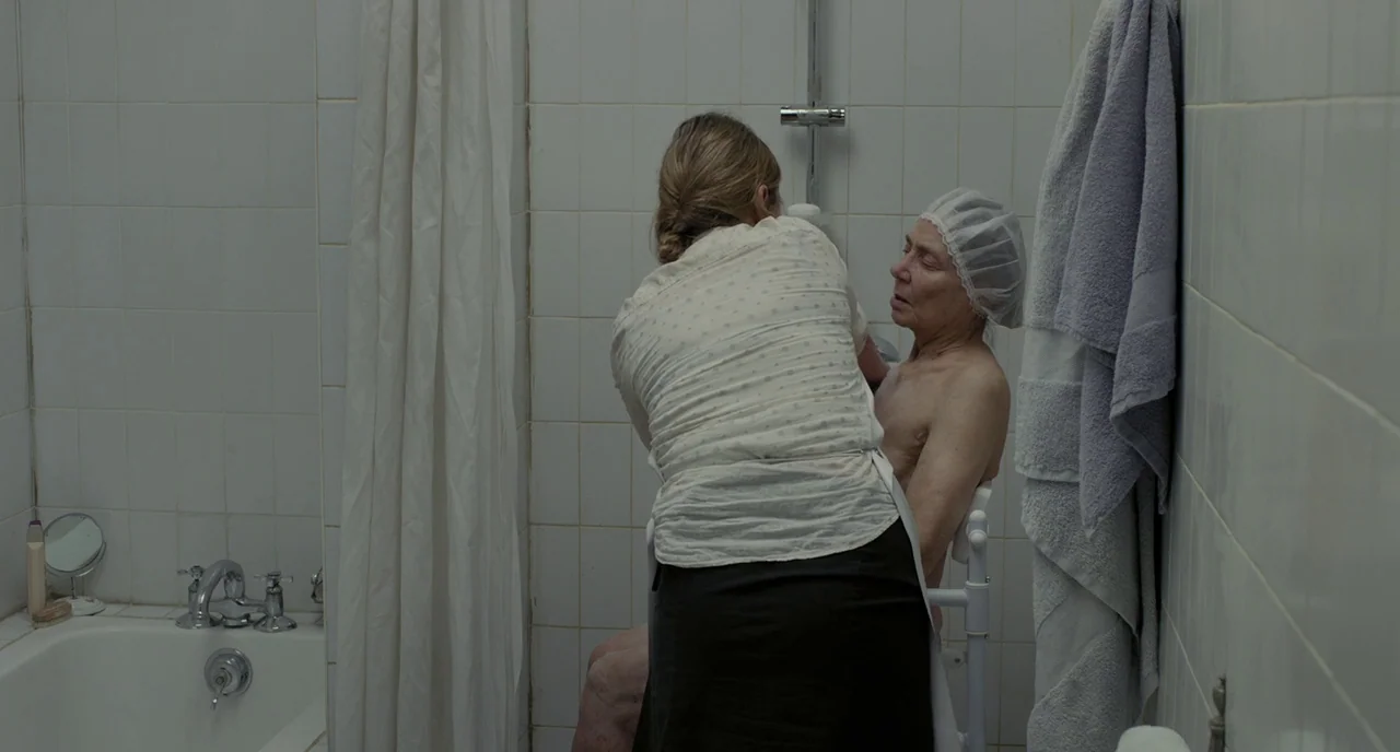 Amour (2012) movie still: medium — A tiled bathroom features a small shower stall and a white porcelain bathtub. A woman assists an elderly person…