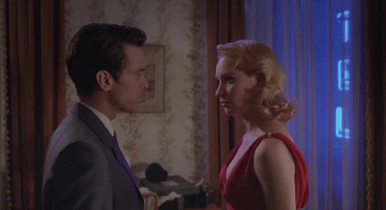 Populaire (2012) movie still: medium — A hotel room features vintage floral wallpaper and a blue neon sign outside the window. A man in a suit and a…