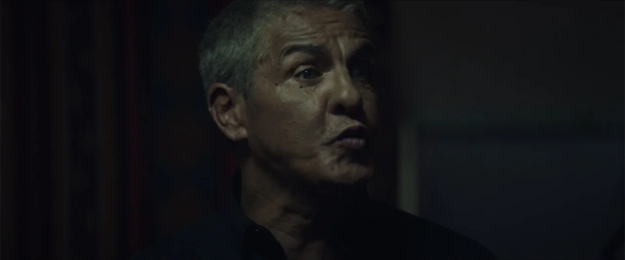 Redemption Day (2021) movie still: closeup — A dimly lit indoor space with dark, indistinct surroundings behind a person. A middle-aged man is…