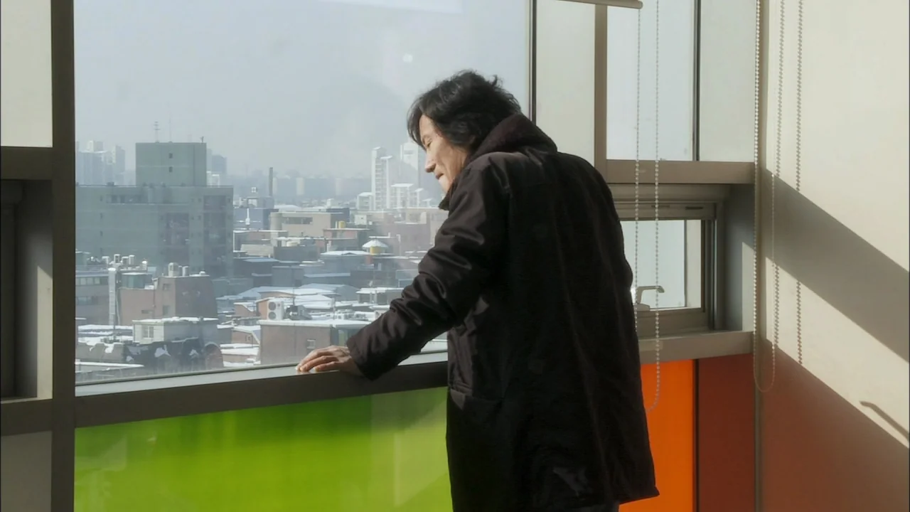 Oki’s Movie (2010) movie still: medium — A high-rise room overlooks a snowy urban skyline on a bright winter day. An elderly man stands by the…
