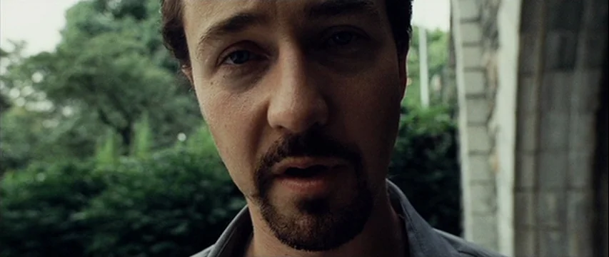 25th Hour (2002) movie still: closeup — A man stands beneath a stone archway with dense green foliage in the background. He is positioned close to…