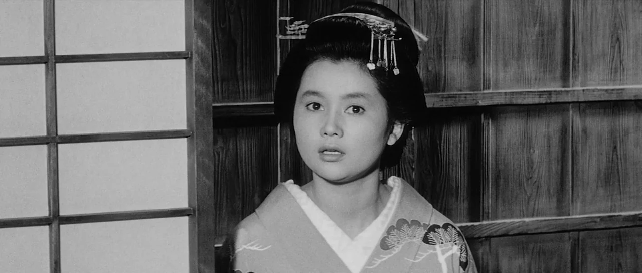 Sanjuro (1962) movie still: medium — A woman stands indoors against a dark wooden wall and a shoji screen. She looks directly toward the viewer with…