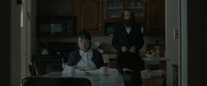 To Dust - Nightmares (2018) movie still: medium — A dimly lit kitchen features wooden cabinetry and a small dining table in foreground. An older…