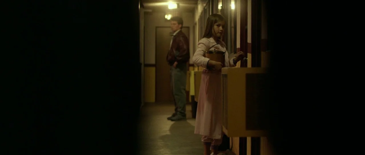 Faults (2014) movie still: fullbody — A dimly lit motel corridor at night has yellow walls and multiple doors. A young girl in pajamas turns a door…