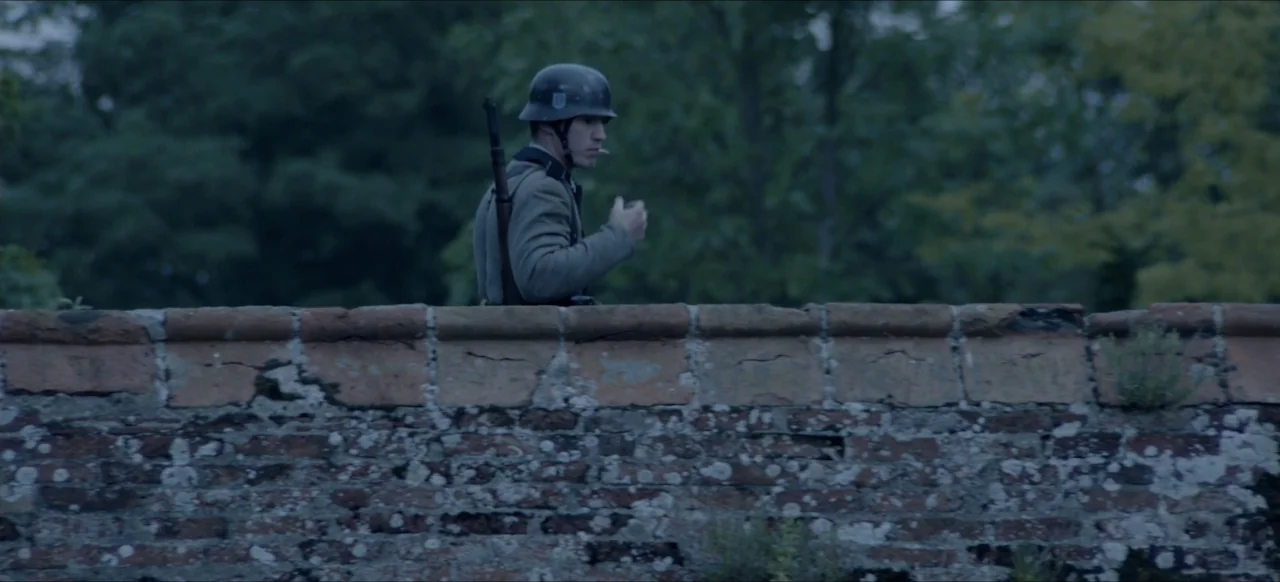 We Go In at Dawn (2020) movie still: medium — A soldier in a uniform stands behind a textured moss-covered brick wall outdoors. The man holds a lit…