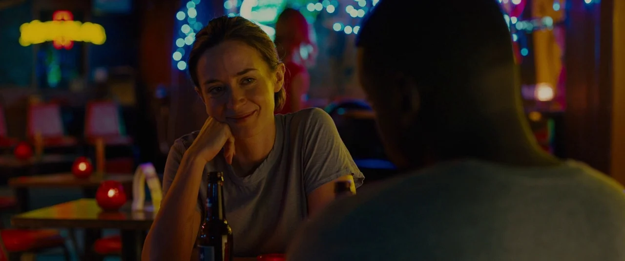 Sicario (2015) movie still: medium — A dark bar setting with red candles on wooden tables and blurred neon lights. A woman in a grey shirt smiles…