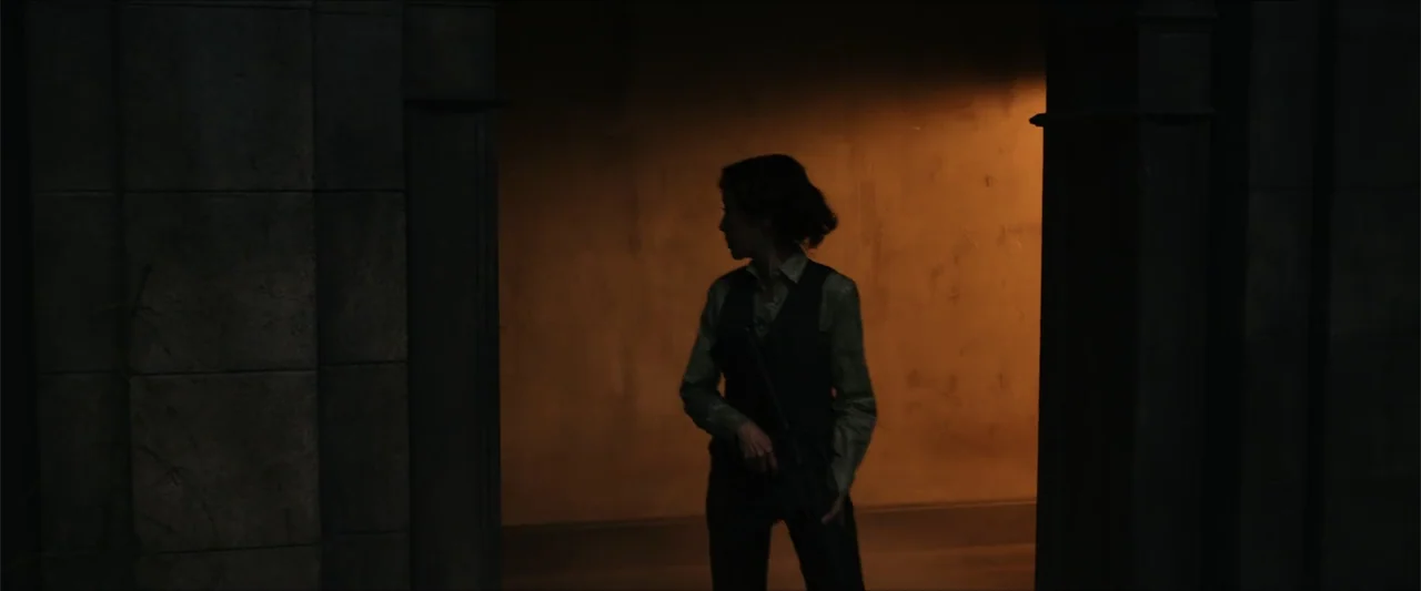 Gunpowder Milkshake (2021) movie still: medium — A dark stone-walled corridor transitions into a brightly lit amber space. A woman stands in the…