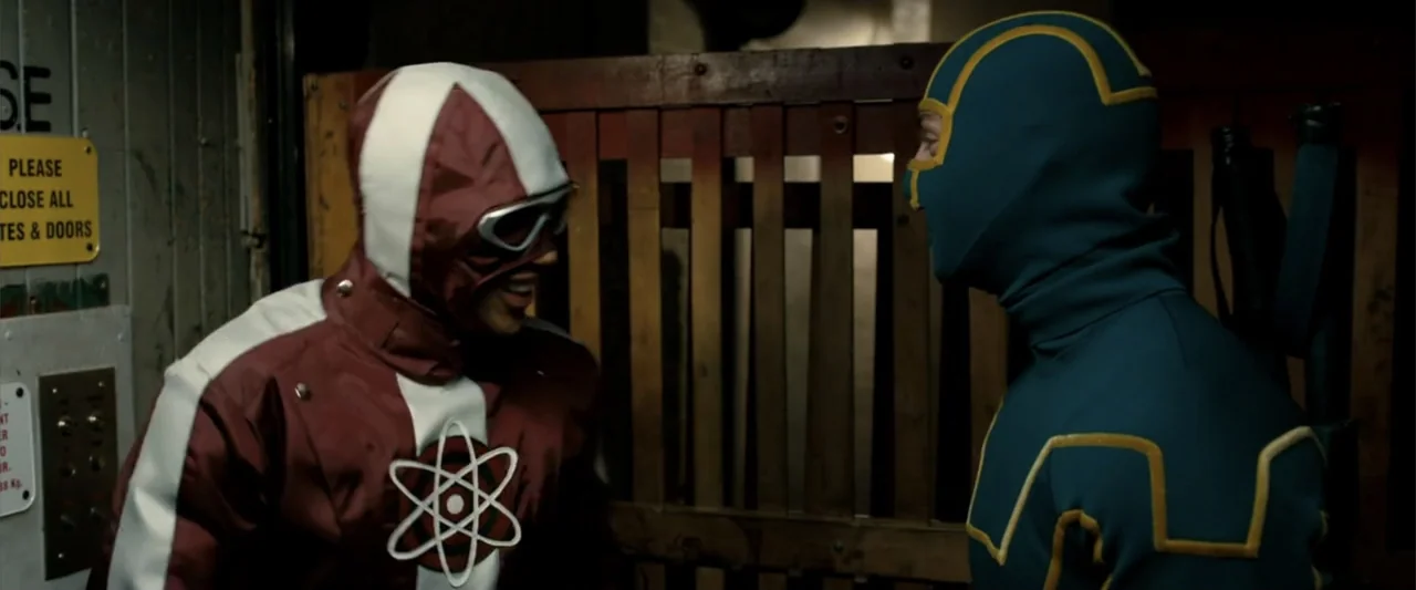 Kick-Ass 2 (2013) movie still: medium — A dimly lit indoor passage features a brown slatted wooden gate and utility panels. Two men in superhero…