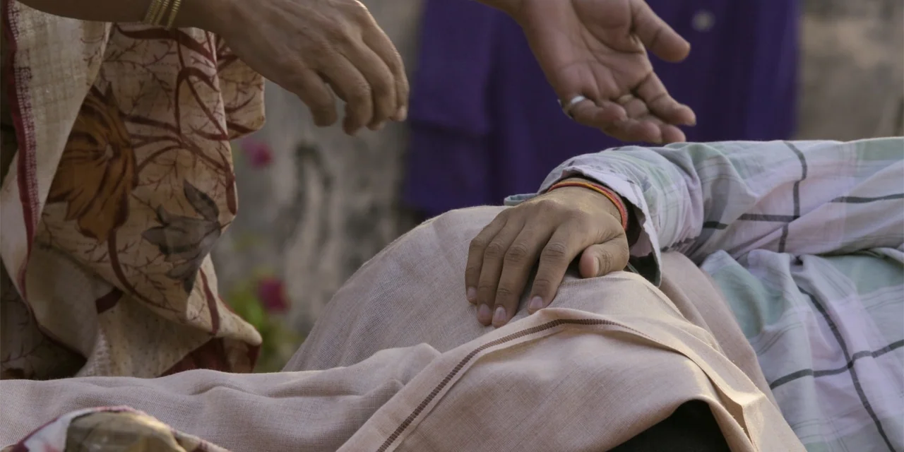 Dry Day (2023) movie still: closeup — An outdoor area with blurred foliage and a stone wall in the background. One person reaches out a hand toward…