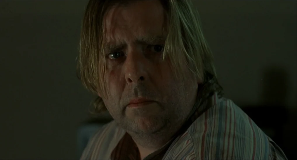 All or Nothing (2002) movie still: closeup — The subject stands in a dimly lit, nondescript indoor space with olive-grey walls. A man with thinning…