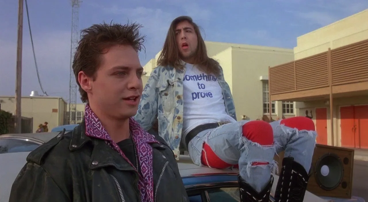 Heathers movie still: medium — A paved lot in front of a tan commercial building with red double doors. Two young men in 1980s fashion linger near a…