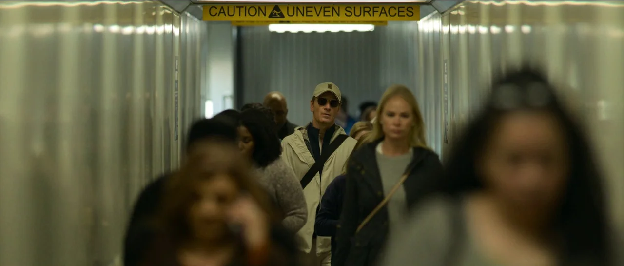 The Killer (2023) movie still: medium — An enclosed passenger jet bridge with metal side walls and a warning sign above. A man in a baseball cap…