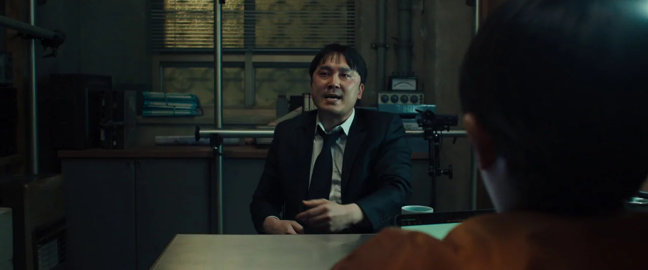 Decision To Leave (2022) movie still: medium — A dimly lit room with metal filing cabinets and electronic equipment along the back wall. A man in a…