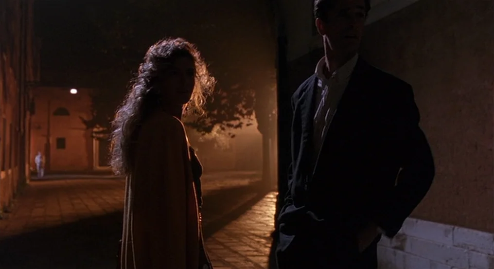 The Comfort of Strangers (1990) movie still: medium — A dark urban alleyway features stone pavement and amber light from an unseen lamp. A man and…