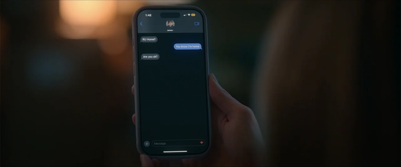 Love Me Love Me (2026) movie still: closeup — A person holds a dark smartphone displaying an iMessage conversation against a blurred background. The…