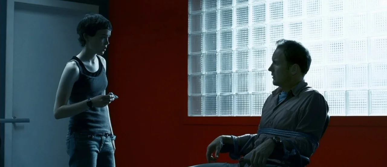 Hard Candy (2005) movie still: medium — A modern room features a striking red wall and a large window with glass bricks. A woman stands in front of…