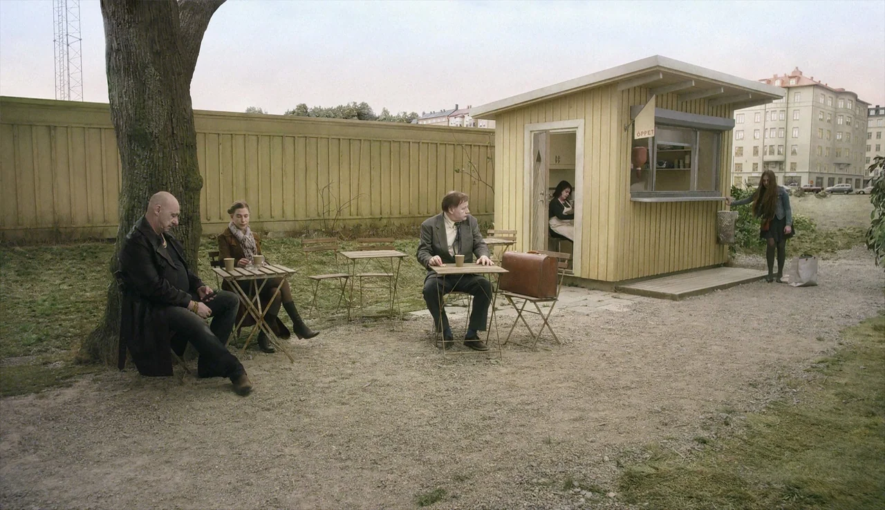 A Pigeon Sat on a Branch Reflecting on Existence (2014) movie still: wide — A gravel-covered open area features a yellow kiosk, a large tree, and…