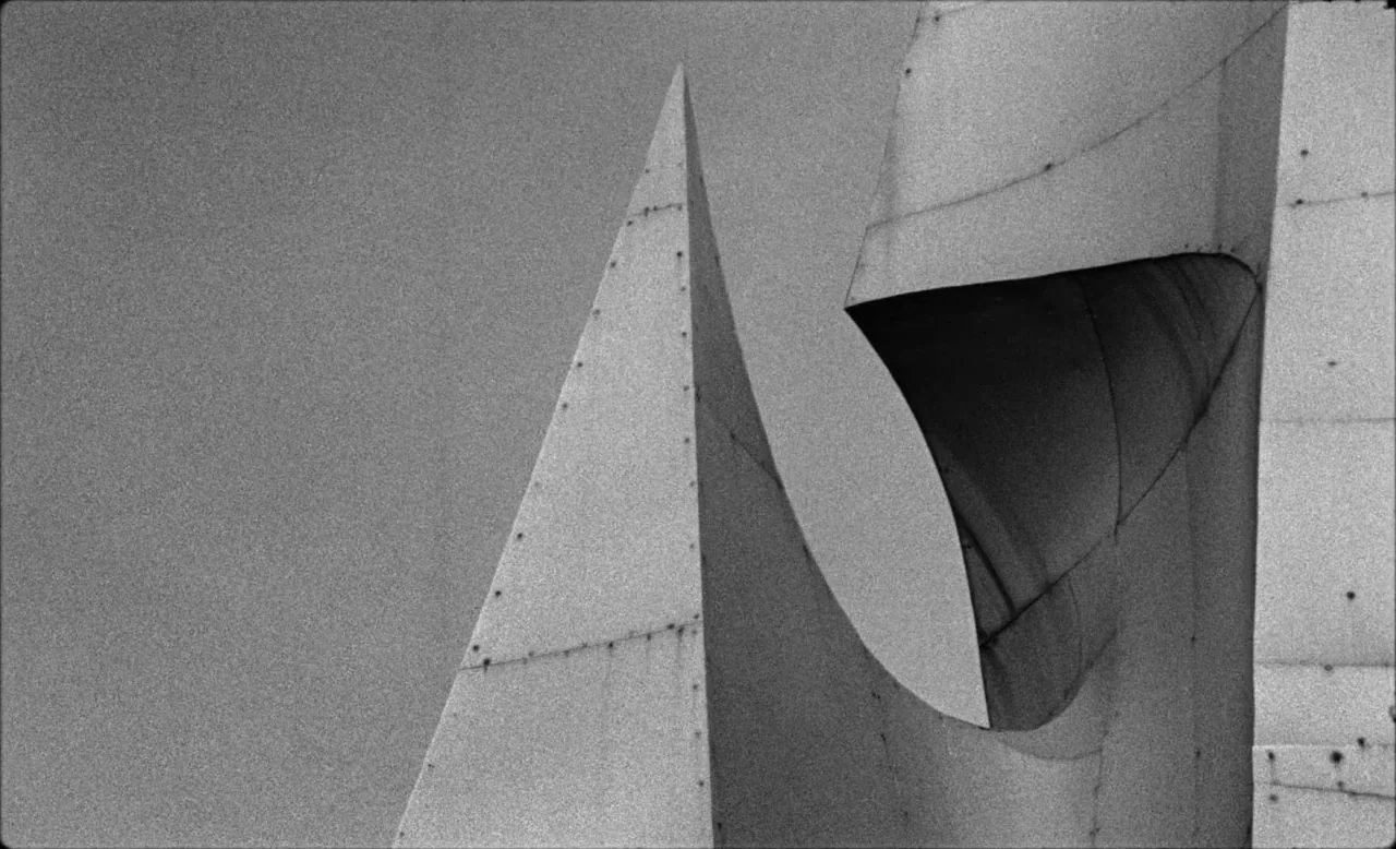 Last and First Men (2020) movie still: closeup — Sharp angular segments of a concrete monument stand against a featureless overcast sky. The…