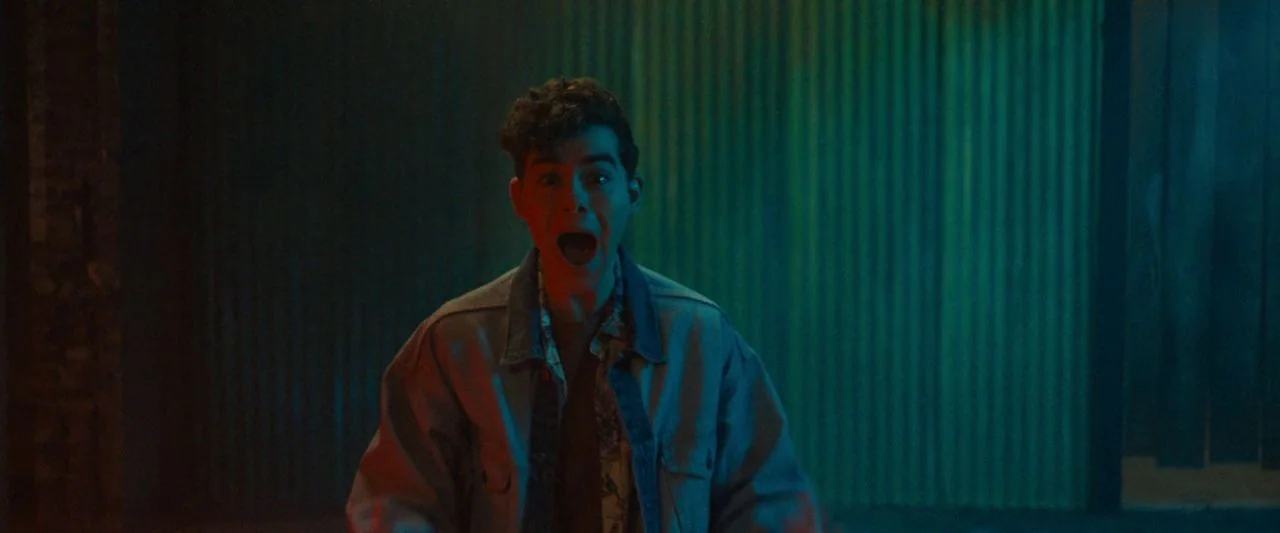 Freaky (2020) movie still: medium — A dark space features walls of corrugated metal illuminated by saturated teal and red light. A young man stands…