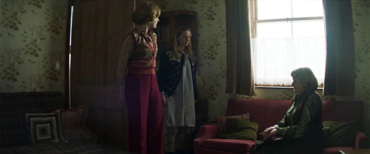 Starve Acre (2023) movie still: fullbody — A dimly lit room features vintage floral wallpaper, a red sofa, and period furniture. Three women occupy…