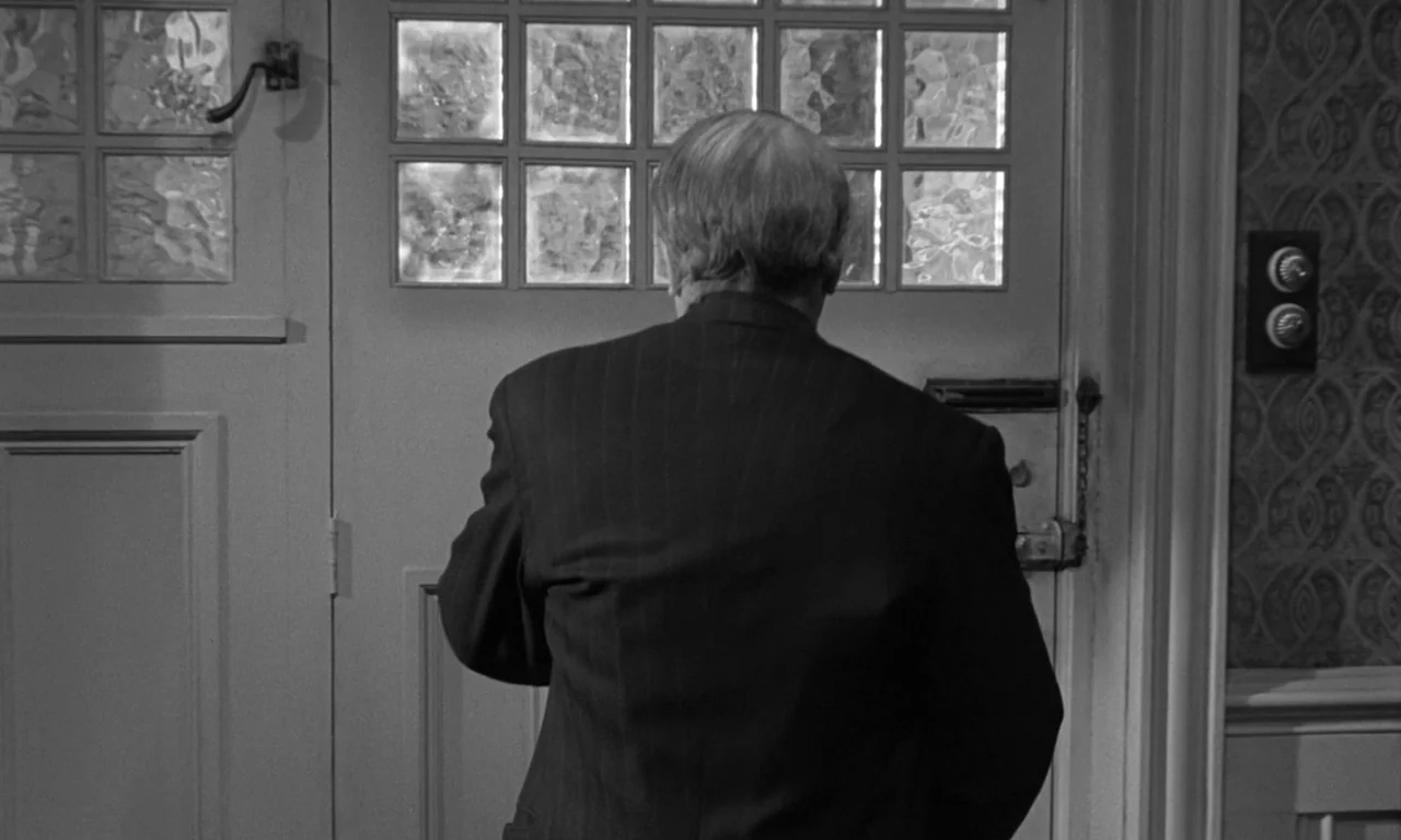 Seance on a Wet Afternoon (1964) movie still: medium — An interior entryway features a wooden door with grid-patterned textured glass windows. A man…