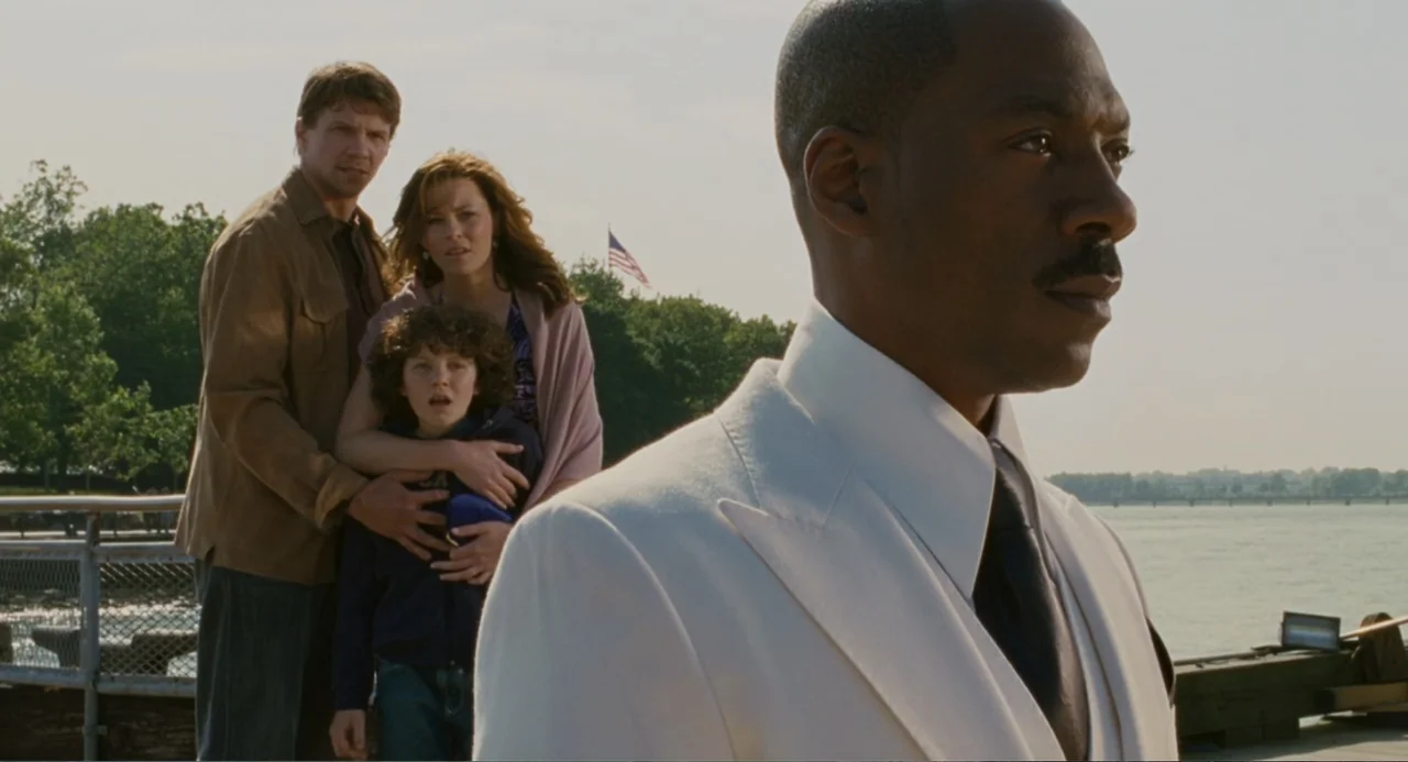 Meet Dave (2008) movie still: medium — A waterfront promenade with a metal railing and a distant American flag visible. A man in a white suit stands…