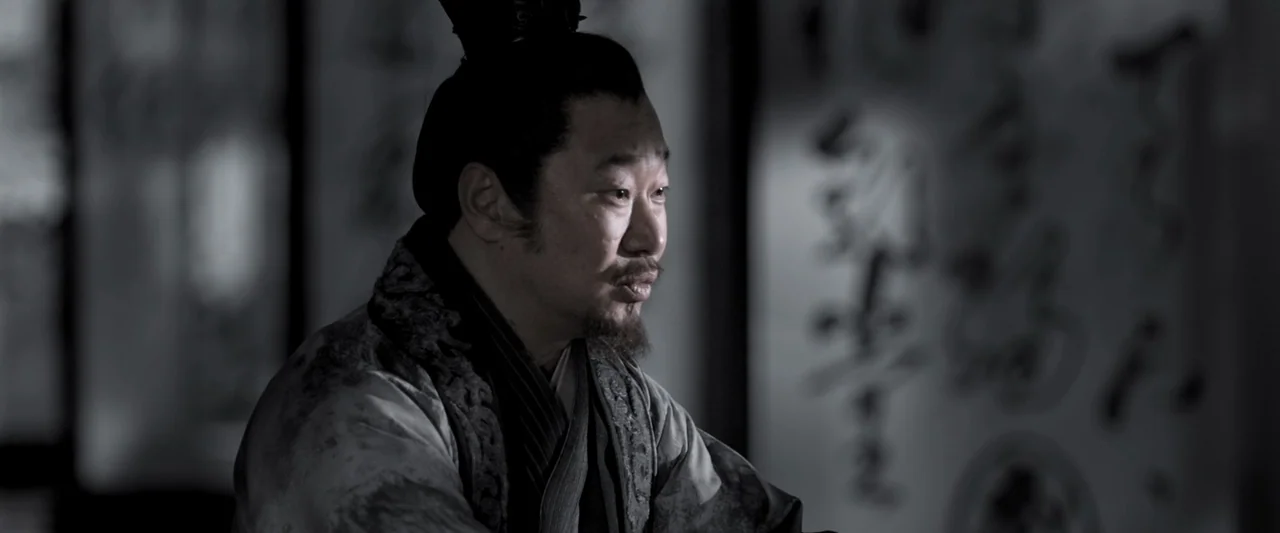 Shadow (2018) movie still: medium — An interior space with a blurred scroll featuring black ink calligraphy hanging in background. A middle-aged man…
