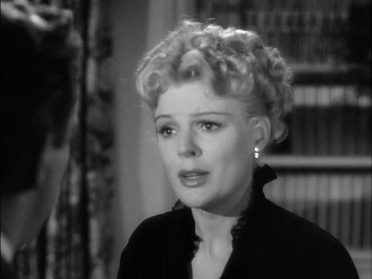 D.O.A. (1949) movie still: closeup — A woman stands in a dim library with bookshelves visible in the blurred background. She turns her head slightly…