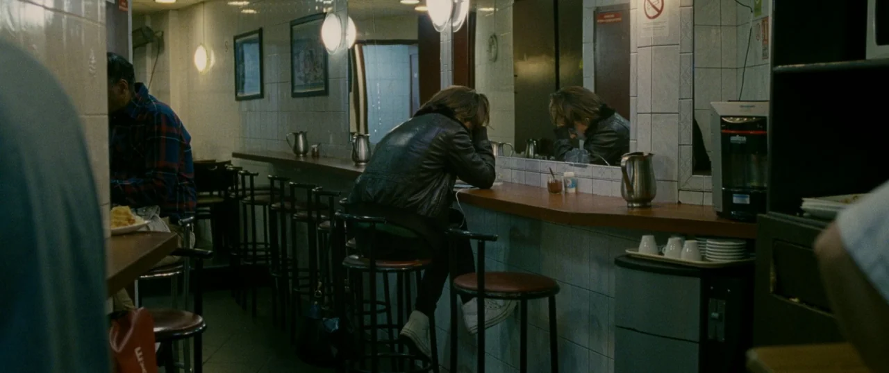 Personal Shopper (2016) movie still: medium — A tiled cafe interior with a long wooden counter and circular metal stools. A person wearing a leather…