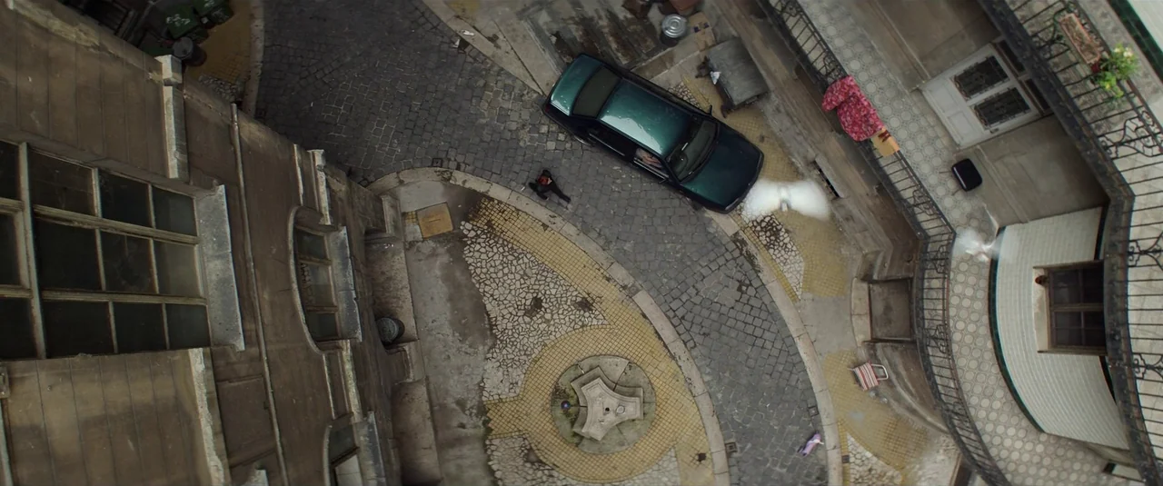 Black Widow (2021) movie still: establishing — A top-down view shows a stone courtyard with a central fountain and dark car. Two white pigeons glide…