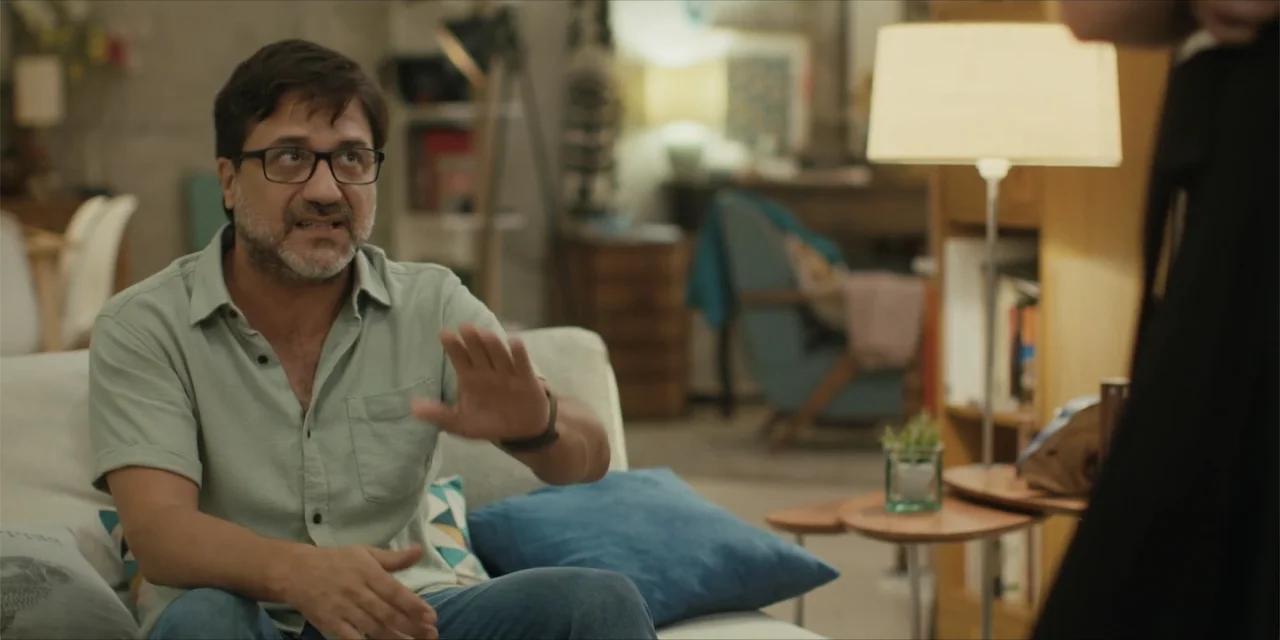 Instant Love (2020) movie still: medium — A furnished living room features a sofa, a floor lamp, and scattered bookshelves. A middle-aged man with…