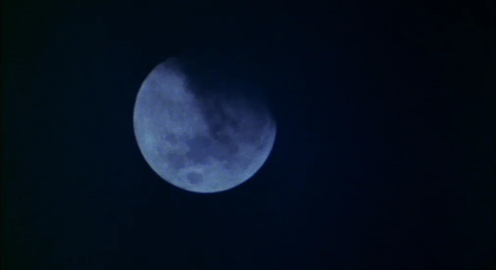 An American Werewolf in London (1981) movie still: closeup — A dark night sky frames a partially clouded, illuminated lunar disc. Thin wisps of…