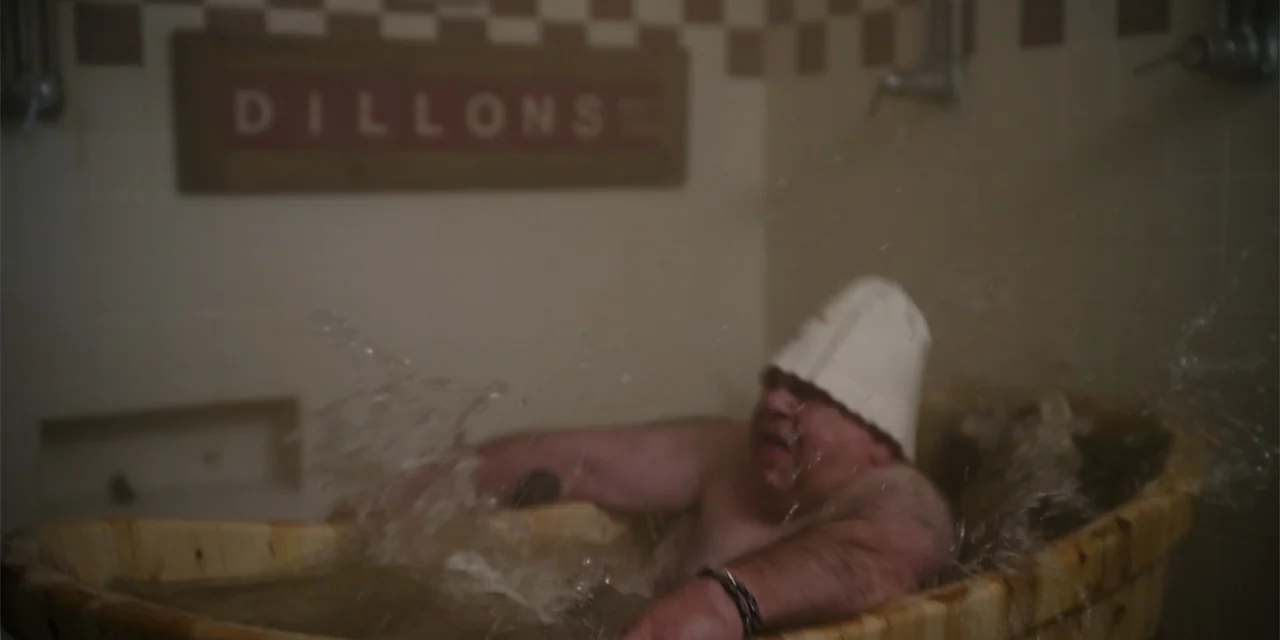 About Fate (2022) movie still: medium — A bathroom with checkered wall tiles and a large wooden soaking tub. An overweight man with a tattoo on his…