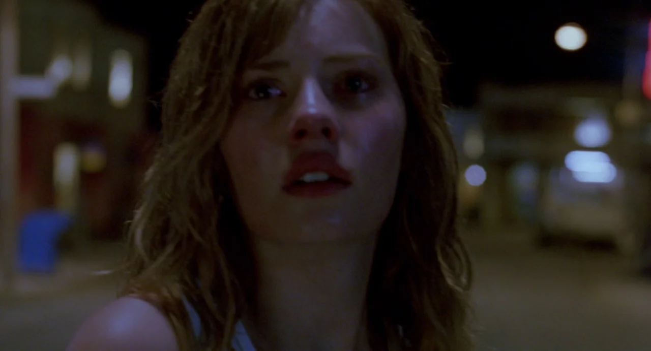 House of Wax (2005) movie still: closeup — A dark city street at night with indistinct blurred structures in the background. A young woman looks…
