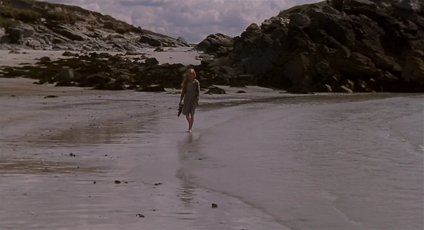 The Secret Of Roan Inish (1994) movie still: wide — A secluded coastal beach framed by dark, craggy rock formations under a cloudy sky. A young girl…