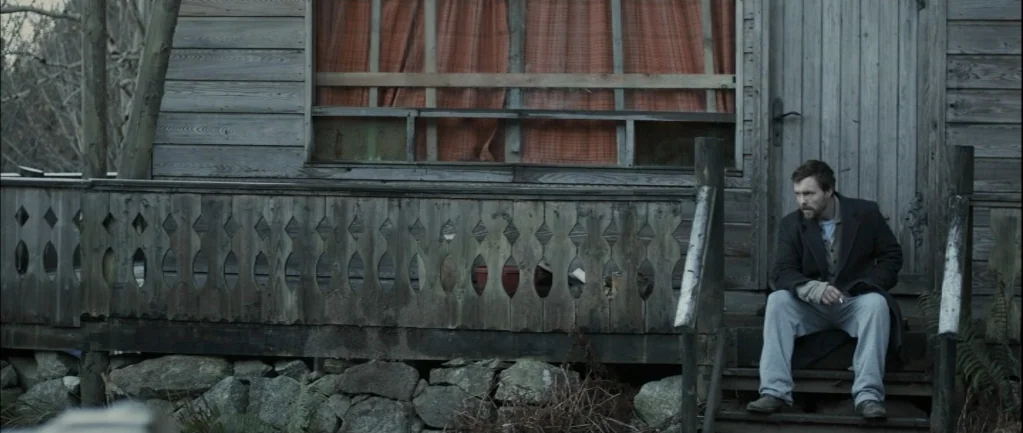 One Hundred Mornings (2009) movie still: medium — A weathered wooden house with a decorative railing and an orange curtained window. A man sits…