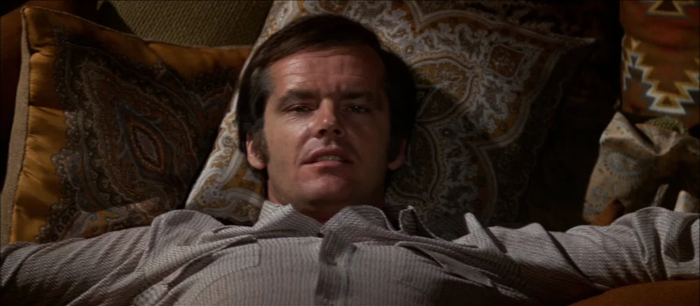Carnal Knowledge (1971) movie still: medium — A man rests against several patterned cushions in a dim residential interior space. He lies on his…
