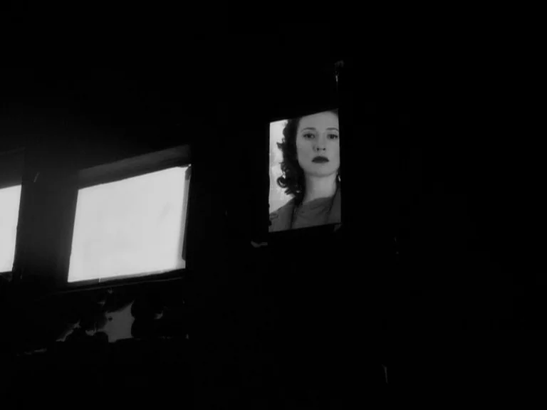 The Good German movie still: closeup — A dark movie theater interior shows a woman's face appearing on a lit screen. The surrounding theater…