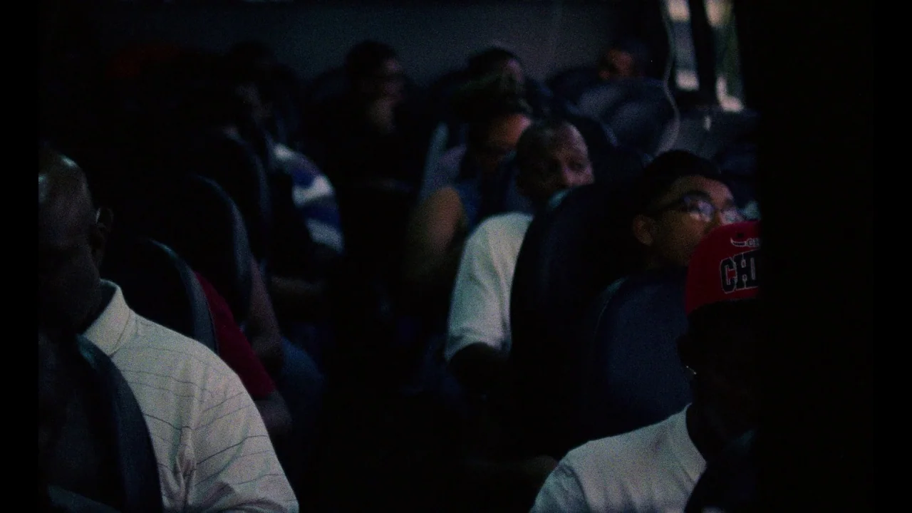 Pass Over (2018) movie still: medium — A dimly lit passenger cabin of a bus is filled with several people seated. Passengers occupy rows of…
